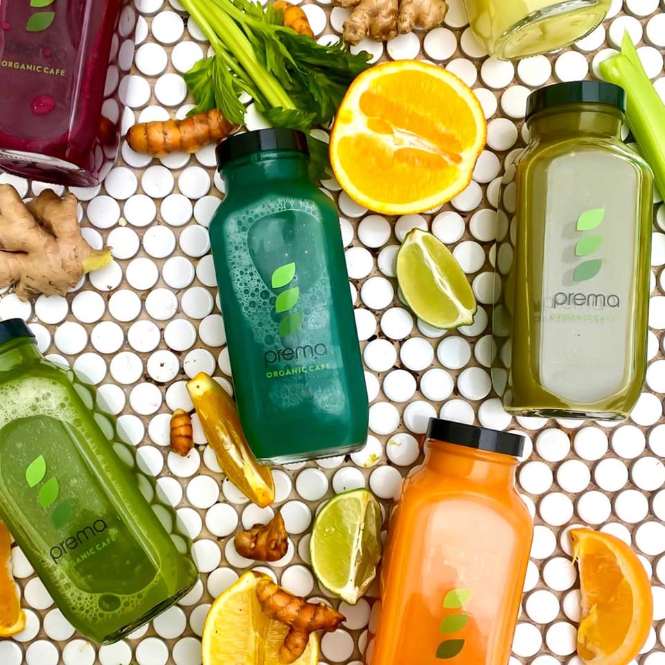 Five Prema Organic Cafe juice bottles in various colors, surrounded by fresh fruits and vegetables.