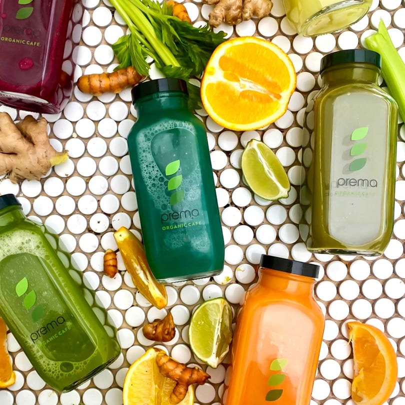 Five Prema Organic Cafe juice bottles in various colors, surrounded by fresh fruits and vegetables.