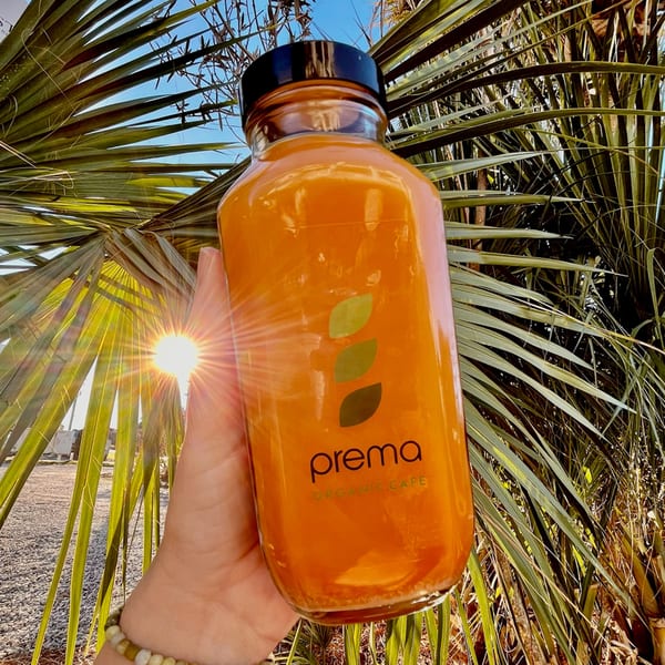 Hand holding a Prema Organic Cafe juice bottle with palm leaves and sun in the background.