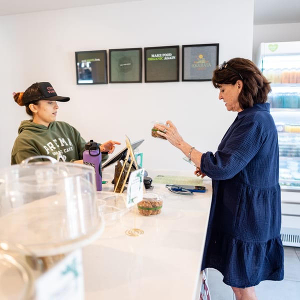 A customer in a blue dress receives a packaged meal from a Prema Organic Cafe employee.