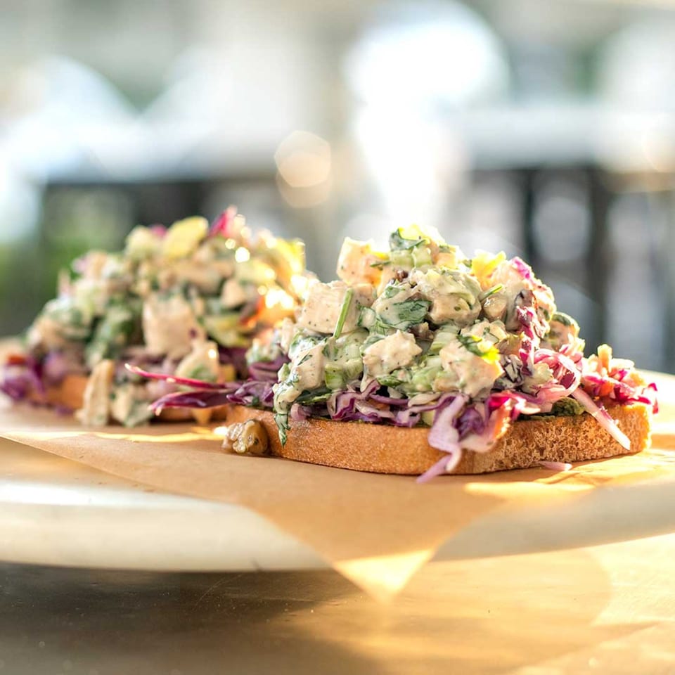 Two open-faced chicken salad sandwiches with red cabbage on brown paper, bathed in warm sunlight.