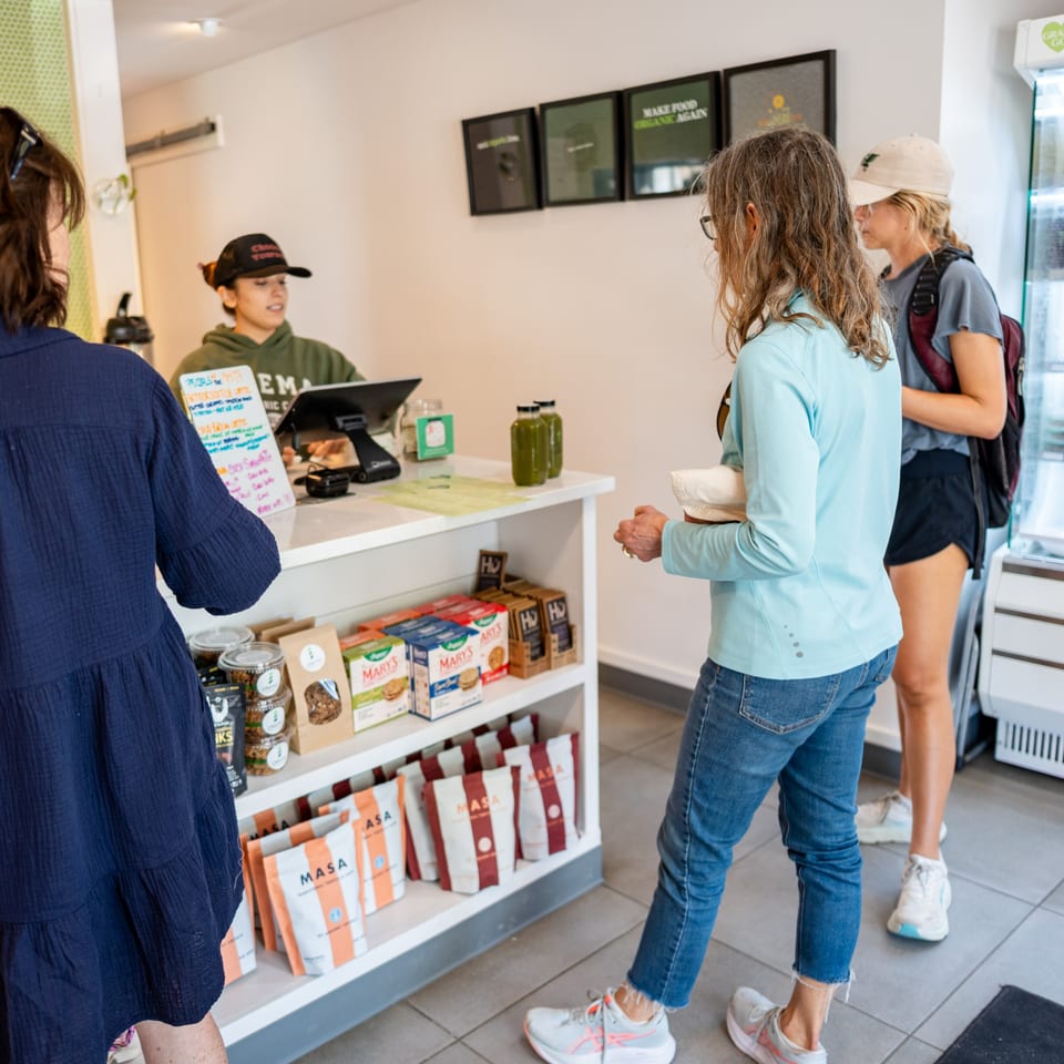 Customers wait in line at a counter with a cashier, shelves of products, and green juice bottles.