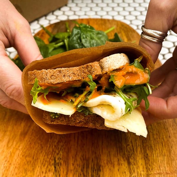 Hands holding a sandwich with cheese, arugula, and orange sauce, wrapped in brown paper.