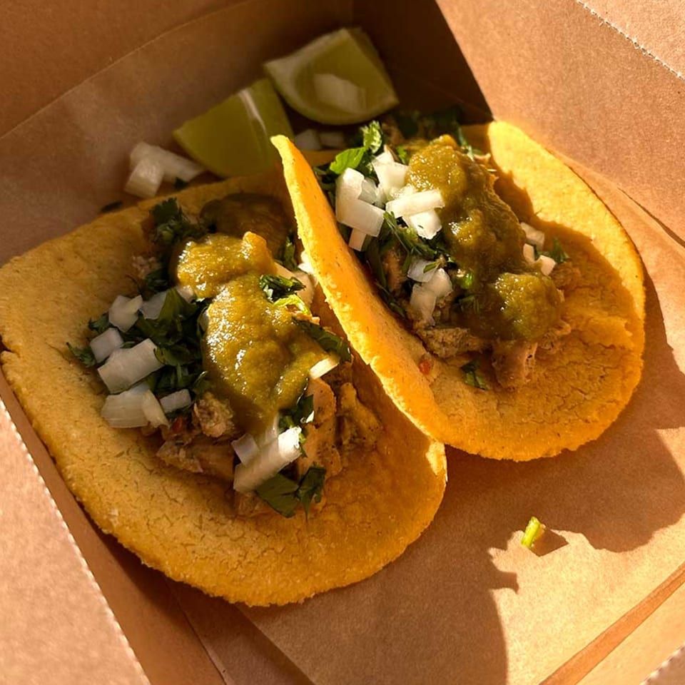 Two tacos with green salsa, diced onions, and cilantro in a brown takeout box with lime wedges.