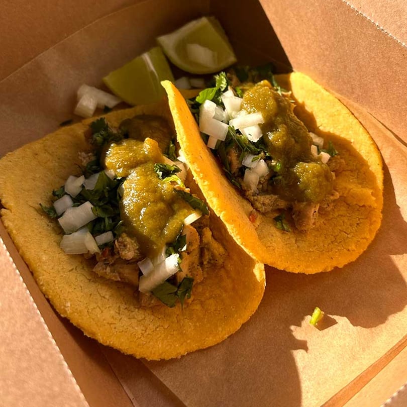 Two tacos with green salsa, diced onions, and cilantro in a brown takeout box with lime wedges.