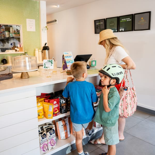 A woman and two young boys, one in a helmet, stand at a cafe counter with snacks.