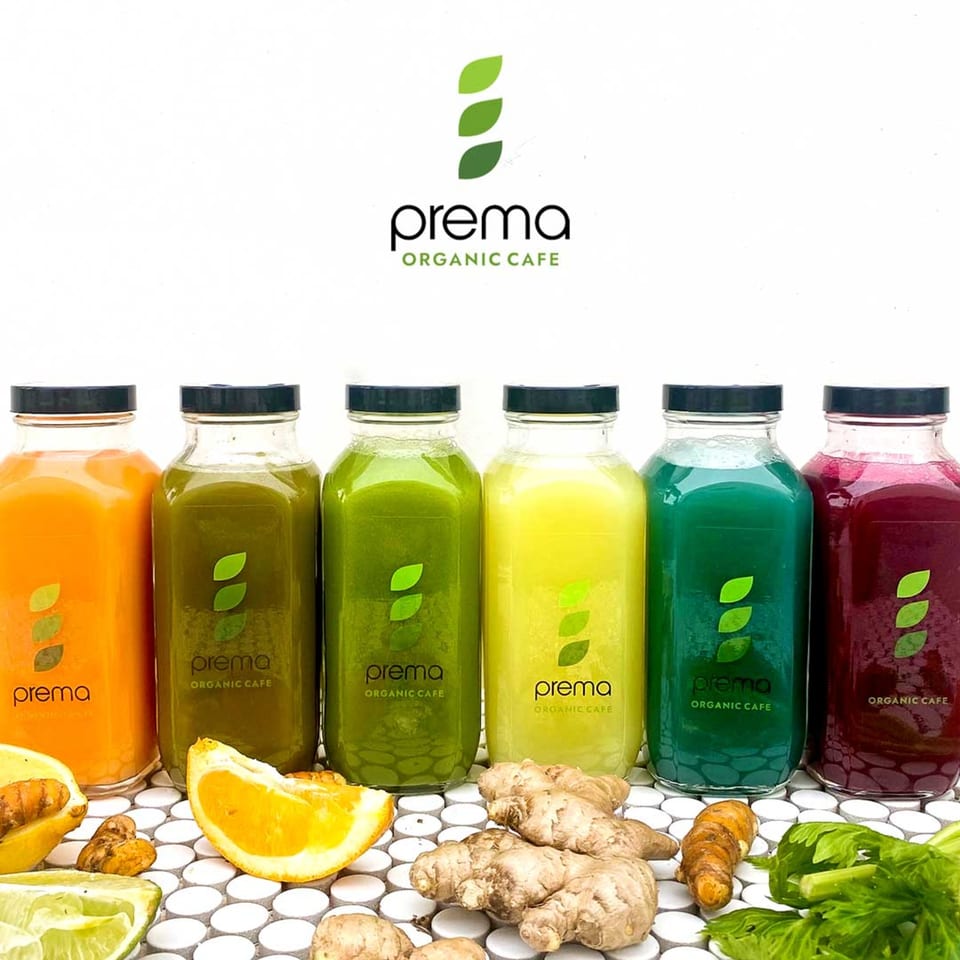 Six colorful Prema Organic Cafe juice bottles with fresh ginger, turmeric, orange, and lime.