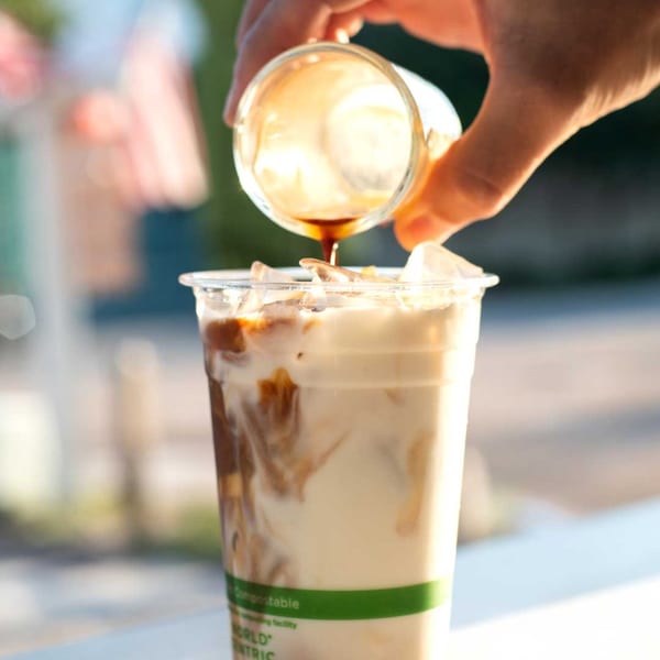 A hand pours espresso into an iced latte in a clear, compostable cup.
